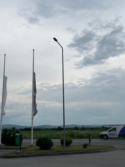 The image captures an outdoor scene on an overcast day, possibly in Hălchiu, Romania. The sky is filled with dramatic, grey clouds, suggesting a moody or potentially rainy atmosphere. In the foreground, several flagpoles stand vertically. Two flags are partially visible; one is dark with a logo and the other is lighter. A tall street lamp with a curved arm extends into the frame from the top left.
Further into the background, a paved area transitions to a grassy verge with bushes and trees. Beyond that, a road is visible with a white van. The van has the "DIGI" logo printed on its side, along with some smaller text that is difficult to read. The van appears to be stationary on the road. In the distance, rolling hills and mountains are silhouetted against the cloudy sky, indicating a rural or semi-rural location. A green trash bin is situated on the grassy verge near the road. The overall impression is one of a functional, possibly industrial or commercial area adjacent to a natural landscape. The time of day is indeterminate due to the heavy cloud cover.