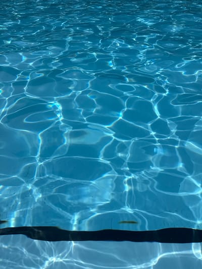 The image displays the surface of a swimming pool, characterized by the dynamic interplay of light and water. The water is a clear, bright blue, with rippling patterns and light refractions creating an abstract mosaic of bright highlights and darker shadows across the surface. These patterns suggest the movement of water, likely due to light interference or slight waves.

On the right side of the frame, a dark, vertical band runs along the edge of the pool. This appears to be a dark coping stone or a line marker within the pool. The texture of the pool floor is faintly visible through the water in some areas, particularly towards the right, where a grid-like pattern can be discerned beneath the shimmering surface.

There are no people or animals visible in the image, nor any overt signs of a specific activity beyond the natural movement of water. The lighting conditions suggest it is daytime, with sunlight creating the strong refractions. The clear blue of the water and the bright highlights imply fair weather. The presence of a swimming pool itself indicates an outdoor recreational setting, possibly a residential or public pool. The image does not contain any visible text. The context of Saint-Imier, Switzerland, suggests this is a scene from that geographical location.