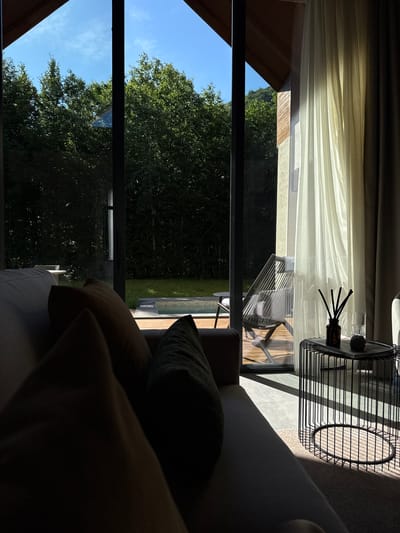 The image shows a view from a modern living space looking out through large glass doors to a natural outdoor environment. In the foreground, a portion of a sofa with dark cushions is visible, partially silhouetted by the strong sunlight streaming in. Adjacent to the sofa, a black wire side table holds a glass of water, a small decorative stone, and a diffuser with reeds in a dark bottle.
Beyond the glass doors, a wooden deck leads to a small, partially visible swimming pool. A lounge chair is placed on the deck. The outdoor area is surrounded by lush green trees and a grassy lawn. The sky is a clear, bright blue. The architecture of the building features angled wooden roof beams and concrete or stone walls. The lighting suggests it is daytime, with dramatic shadows cast by the sunlight. The overall scene evokes a sense of modern comfort integrated with a natural landscape, likely in a resort or private residence. The location is described as Bakovići, Montenegro.