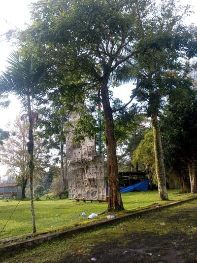 The image shows a recreational area in Mya Sit Kaing Ywar Thit, Myanmar. A tall artificial rock-climbing wall stands in the background, partially obscured by trees. The ground is covered in grass, with scattered litter. Several trees are present, including a palm tree on the left. In the background, there's a structure covered by a blue tarp. A moss-covered concrete border runs along the bottom of the frame. No people are visible.