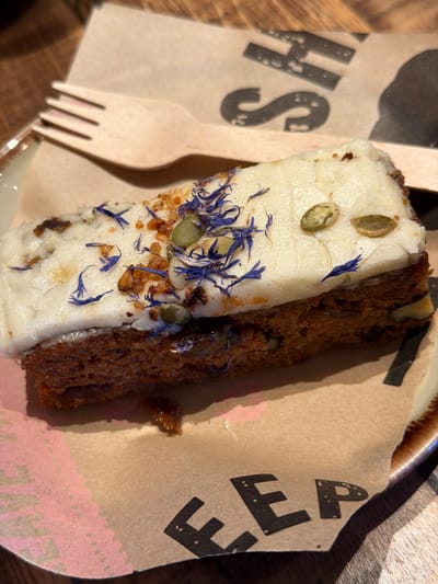The image displays a close-up, static view of a rectangular slice of carrot cake with a thick layer of white cream cheese frosting, served on a crumpled piece of light brown paper. The frosting is garnished with scattered blue cornflower petals, green pumpkin seeds, and small, light brown nut fragments. A light-colored wooden four-tined fork rests on the paper to the upper left of the cake. The brown paper features partially visible inverted black text, displaying "SH" in the upper right portion and "EEP" in the lower left portion, along with a faint pink smudge. The paper rests on a ceramic plate with an irregular speckled brown rim, which is placed on a dark, possibly wooden, table surface. No people are visible. The scene is located in Bretton, United Kingdom.