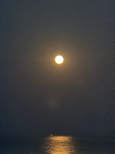 A photograph taken at night in Νέα Καλλικράτεια, Greece, depicts a moonlit scene over a body of water. The moon, a bright, luminous orb, is positioned centrally in the upper portion of the frame, casting a yellowish glow. Its light reflects onto the water below, creating a shimmering, golden path. In the distance, two small, bright lights are visible on the water, likely from a boat or buoy. The overall atmosphere is one of serene darkness, with the moon and its reflection being the primary sources of illumination. The water appears calm, with gentle ripples visible in the reflected moonlight. The sky is dark and hazy, with a subtle texture.