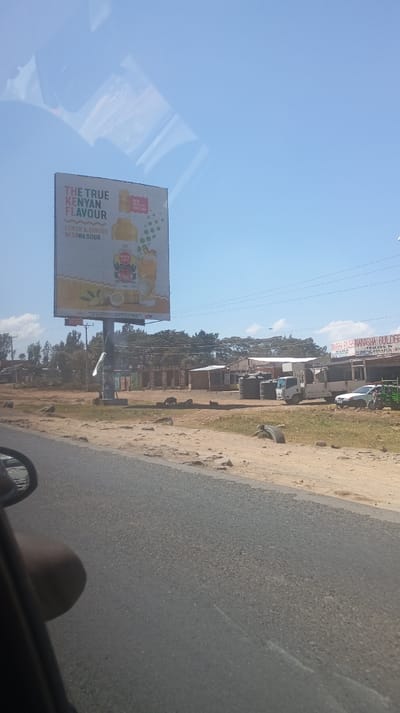 A roadside scene shows a large billboard advertising "The True Kenyan Flavour" with a lemon and ginger drink. A collection of small shops and buildings lines the opposite side of the road. Water tanks, vehicles, and a tractor are visible near the shops. The sky is a clear blue, and the view is taken from inside a car, showing the dashboard and side mirror. The foreground features a paved road with adjacent dirt patches.