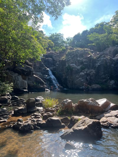 A serene natural scene unfolds with a waterfall cascading down a rocky cliff face into a clear, dark pool of water. Lush green trees and foliage frame the top and sides of the image, with sunlight dappling through the leaves and reflecting on the water's surface. Several individuals are present in and around the water, appearing to be enjoying the refreshing environment. Two people are seated on rocks to the right of the waterfall, while others are visible in the water near the falls, possibly swimming or wading. The rocky terrain extends into the foreground, with various sized rocks scattered along the water's edge. The overall impression is one of a tranquil, natural oasis, potentially a popular spot for recreation and relaxation. The location is identified as Vĩnh Hải, Vietnam.