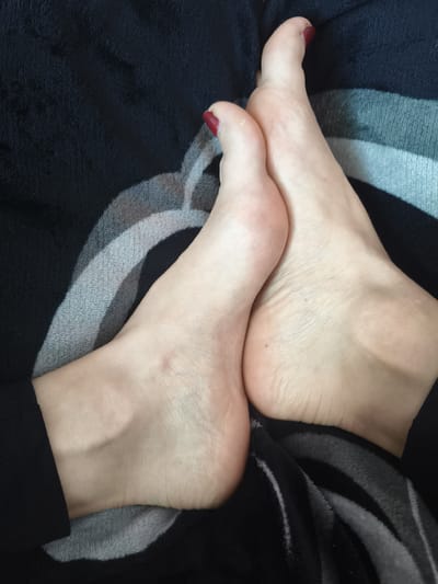 A close-up, overhead shot shows two bare feet positioned side-by-side against a dark, plush blanket with a gray and white striped pattern. The feet are light-skinned and appear to be from the same individual. The toenails on both feet are painted a dark red color. The left foot is angled slightly upwards, revealing the sole and arch, while the right foot is positioned more flatly, with its sole facing towards the camera. The skin on the soles of the feet shows some texture and creases. The background is dominated by the soft fabric of the blanket, which is draped and folded. The image is lit from above, casting subtle shadows on the feet. There are no other people or objects visible in the frame. The scene is domestic and appears to be indoors.