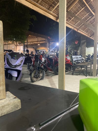 The image captures a nighttime scene, likely an outdoor or semi-outdoor parking area at a restaurant or communal space in Gampongaceh, Indonesia. The perspective is slightly elevated, looking down.

In the immediate foreground, a dark, flat surface, possibly a table, is visible on the right, with a bright green object partially seen at the top right, suggesting the vantage point of the camera. Prominent wooden beams stretch across the frame, dividing the view.

The main subjects are numerous motorcycles and scooters parked side-by-side. In the foreground, a distinct purple/lavender scooter is parked, its headlights visible. Behind it, several red and black motorcycles/scooters are lined up.

To the left and top of the frame, a traditional thatched roof structure with visible wooden supports extends, indicating a rustic architectural style. Beyond the parked vehicles, artificial lights illuminate the area, creating lens flares, and reveal more distant motorcycles and a railing made of wood and metal. Dark, leafy trees and bushes are visible in the dimly lit background.

The overall impression is a bustling evening spot, characterized by the presence of multiple vehicles and local architecture. No discernible human activity or legible text is visible.