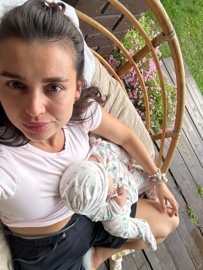 In Sylva, Russia, a woman with dark hair tied back in a bun is captured in a selfie while holding a baby. She is wearing a pink t-shirt and black shorts, and the baby is dressed in a white onesie with a floral pattern. They are seated in a round, woven chair on a wooden deck. Pink flowers are visible behind them, and a patch of green grass can be seen to the right. The woman has brown eyes and is looking directly at the camera. The baby's head is nestled against the woman's chest.