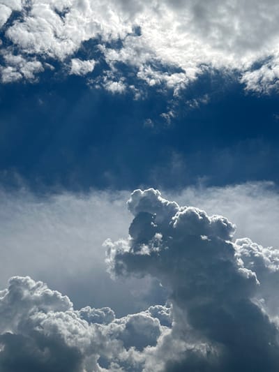 This is a photograph of a dark blue sky filled with dramatic, billowing clouds. The clouds are illuminated from above, with bright white edges and darker undersides, suggesting a powerful light source. In the foreground, large, cumulus clouds dominate the lower half of the frame, casting shadows on each other and creating a sense of depth and volume. Higher in the sky, thinner, more wispy clouds stretch across the frame, interspersed with patches of clear blue sky. The image appears to be taken looking upwards, capturing the dynamic and ever-changing nature of the sky. The scene is devoid of any people, structures, or terrestrial elements, focusing solely on the atmospheric conditions. There is no indication of the city of Pravda, Russia, or any other geographical location.