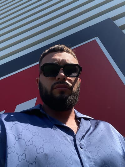 A low-angle, close-up, outdoor, eye-level selfie features a man with dark hair and a full beard wearing sunglasses and a blue button-up shirt with a repeating "GC" pattern. He is positioned in front of a large, red, angular structure with horizontal grey and dark blue stripes above it. The lighting suggests it is daytime, casting a shadow across the man's face. The image appears to have been taken in the city of Korkmaskala, Russia, given the context.