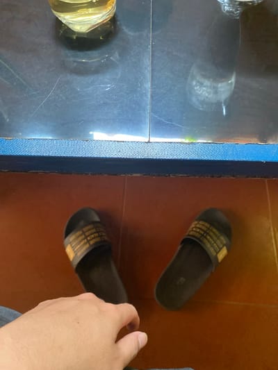 A high-angle shot displays a juxtaposition of surfaces and objects. To the left, a glossy, reflective surface, possibly glass or polished stone, reflects light and what appears to be the base of a glass container with a yellow liquid. A blue textured border runs vertically, dividing this surface from another area. To the right of the border, a brown tiled floor is visible, upon which two black sandals with patterned straps are placed. A hand is partially visible on the right side, seemingly reaching for or touching one of the sandals. The environment suggests an indoor setting, possibly a home or building interior. The location is Đak Đoa, Vietnam.