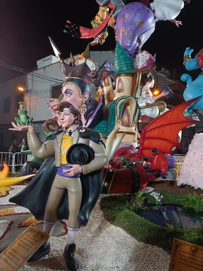 The scene depicts a large, multi-component artistic installation, likely a Fallas monument, at night in El Port de Sagunt, Spain.
In the foreground, a life-sized statue of a male figure, resembling a prince or suitor, stands facing slightly left. He wears a tan jacket over a yellow patterned shirt, a purple sash, tan trousers, black shoes, and a dark cape. His left hand holds a black hat, while his right hand, extended forward, balances a green frog wearing a small gold crown. Behind him, a larger female figure with pale skin, dark hair, and a dark, elaborate crown and dress has her right hand raised near the male figure's head. Her expression appears stern or cunning.
To the right of these figures, a tall, multi-tiered castle structure with teal turrets and golden accents rises. A smaller female figure, dressed in white with intricate patterns, is seated on a section of the castle, holding an open book and illuminated by an unseen light source. Below the castle, a red dragon-like creature with prominent red wings lies on the ground. Various other abstract and figural components are visible higher up and to the far right, including a large purple, patterned orb and part of a light blue genie-like figure. A white, textured conical object is partially visible on the far right.
The ground around the installation is covered in light-colored gravel or small stones, interspersed with patches of artificial green grass, some containing small white and purple flowers, and a small, dark, reflective pool. Two brown plaques with text, presumably descriptions in Valencian or Spanish, are positioned on the ground.
The background shows a dark night sky. A streetlight with an orange glow and the upper portion of a light-colored building are visible in the distance. A metal barrier and faint silhouettes of people are present in the far left background, indicating a public viewing area. Artificial lighting illuminates the monument, casting shadows and highlighting its various colorful elements.