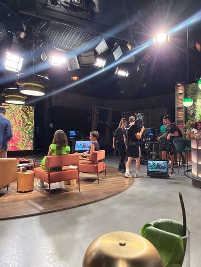 This is an indoor scene, likely a television studio, set in the city of Sant Joan Despí, Spain. The studio features a raised wooden platform with a curved edge, on which two people are seated in orange chairs. A man in a blue blazer stands to the left, facing away from the camera. In the background, a film crew is actively working. One person operates a large video camera on a pedestal, while others are positioned around them, some wearing headsets. Several monitors display video feeds. The studio is equipped with numerous overhead lights, some of which are brightly illuminated, creating lens flares. A large, colorful mural or screen is visible on the left wall. The overall atmosphere suggests a live television production is in progress.