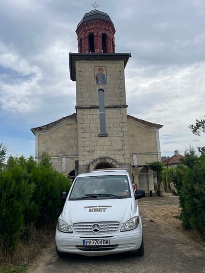 The image depicts a white Mercedes-Benz van parked near the Church of Saint Petka in Tsar Kaloyan, Bulgaria. The church is a stone building with a brick bell tower, adorned with an icon. The sky is cloudy. The vehicle has a Bulgarian license plate, "PP 7704 BM," and text on its windshield is barely visible. The van is adjacent to some greenery.