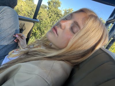 A blonde-haired woman with her eyes closed is reclined in the passenger seat of an open vehicle in Khwai, Botswana. She is wearing a tan jacket and jeans. The vehicle appears to be open-air, with a blue sky and green trees visible in the background.
