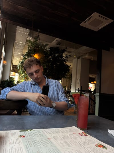 A young man with curly blond hair and closed eyes is sitting at a table, looking down at his phone, which he holds in his right hand. He is wearing a blue and white striped button-down shirt and a watch on his left wrist. To his right, there is a tall glass containing a pink drink with a slice of watermelon and a straw. A menu is placed on the table in front of him, with some text and floral illustrations visible. The setting appears to be a restaurant or bar, with dark wooden beams and paneling on the left, and a bright, airy interior with plants and lighting visible in the background. The overall lighting suggests it might be evening or a dimly lit indoor space. The man's posture and closed eyes could indicate relaxation, tiredness, or deep focus on his phone. There are some faint reflections on the table.