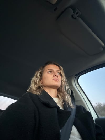 A person with blonde, wavy hair and light-colored eyes looks upward in a car. The person is wearing a white shirt under a dark, textured coat. The car's interior is visible, including a sun visor and a window showing a glimpse of the outside sky. The angle is from below, giving an upward perspective. The setting suggests travel or being a passenger in a vehicle.