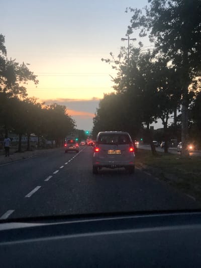 A traffic jam is captured on a road flanked by lush green trees against a vibrant sunset sky. The vehicles, mostly cars and a van with visible license plate "FP 89 36 GP", are lined up in a queue. Dashed white lines mark the lanes on the asphalt road. Streetlights and power lines are also visible, adding to the urban atmosphere. The image seems to be taken from the window of a vehicle.