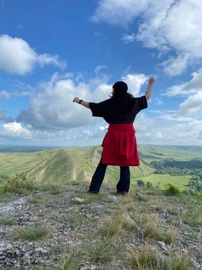 A person wearing a black beanie, black t-shirt, red sweatshirt tied around their waist, and black pants stands on a grassy, rocky hilltop overlooking a valley. The person's arms are raised, with their right arm extended upwards and their left arm bent at the elbow, also raised. They are facing away from the camera, looking towards the valley below. The sky is blue with scattered white clouds. The valley features rolling green hills and a treed area in the distance. The location is stated to be Andreevka, Russia.