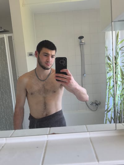 A shirtless man with a beard and a silver chain around his neck is taking a selfie in a bathroom. He is holding a black smartphone. He is partially obscured by a gray towel wrapped around his waist. The bathroom includes a shower with a shower head and faucet, a shower curtain with bamboo leaf patterns, white tiled walls, a mirror, and a white tiled sink. There is no indication of Aystetten, Germany.