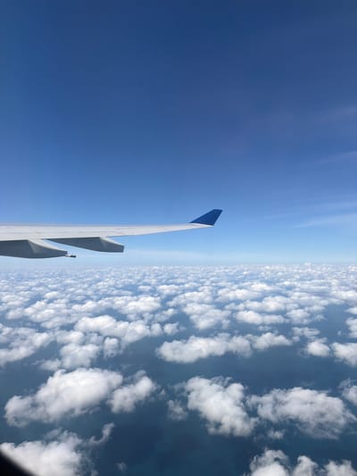 The image shows a view from an airplane window. The left side of the image shows a clear blue sky. The right side shows a dense layer of puffy white clouds extending to the horizon. An airplane wing with a blue wingtip is visible in the foreground, separating the sky and clouds. The image was taken from an aircraft, so the location is likely above a city, but it is not possible to determine the city of Paiania Municipal Unit, Greece from the image alone.