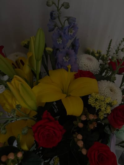 The image displays a close-up, overhead view of a vibrant floral arrangement. The arrangement features several types of flowers in various colors, including bright yellow lilies, deep red roses, and clusters of small white and pale blue flowers, possibly delphiniums. There are also small, light orange berries interspersed among the foliage. The background is a plain, light-colored wall. The lighting is dim, suggesting an indoor setting. The image does not provide any information about the location in the city of Shepetivka, Ukraine, nor does it depict any people or specific actions.