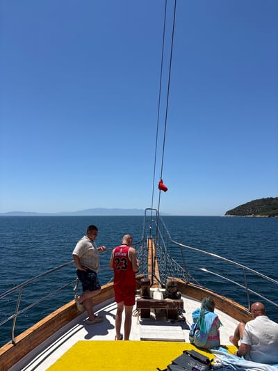 The image captures a sunny day on a boat trip near Menderes, Turkey, with clear blue skies and calm, deep blue water. The photograph is oriented vertically, showing the horizon running up the right side of the frame.

On the wooden deck of the boat, several individuals are present. In the foreground, two men stand. One man, positioned slightly to the right, wears a red basketball jersey with "JORDAN" and the number "23" printed in white. Next to him, another man wears a light-colored striped short-sleeved shirt, patterned shorts, and light-colored clogs. Further back on the deck, an older bald man wearing a white shirt is seated, looking towards the water. Nearby, another person with braided hair is partially visible, wrapped in a blue and pink patterned blanket.

The boat deck features a yellow matting covering part of the surface, contrasting with the natural wood and white sections. Metal railings run along the side of the boat, separating the deck from the sea. Ropes or cables stretch across the frame, with a small red flag or piece of fabric attached to one in the mid-ground.

In the distance, the shoreline of hills or mountains is visible under a bright, cloudless sky, suggesting a coastal area. The strong sunlight casts shadows from the boat's structures and the people. On the deck, blue and white towels are casually placed, and a black bag with "NORTH FACE" visible on it, along with a white cup, are also present. The scene conveys a relaxed, leisurely atmosphere on the water during daytime.