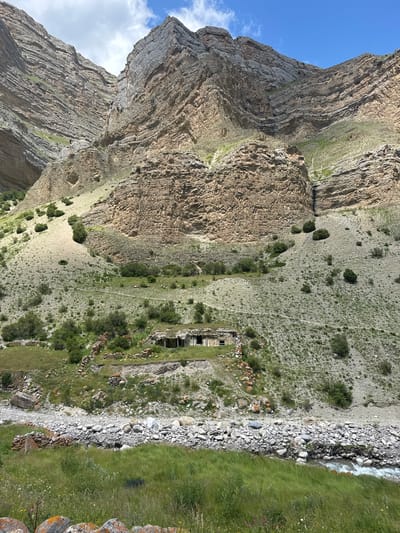 The image depicts a mountainous landscape with a small, dilapidated stone structure built into the hillside. The structure appears to be ancient, with a crumbling roof and visible openings that resemble windows or doorways. The surrounding environment is characterized by steep, rocky slopes covered with sparse vegetation, including grass and shrubs. A clear stream flows in the foreground, its banks lined with smooth stones. The sky above is partly cloudy with a bright blue expanse. The scene suggests a remote and possibly abandoned settlement in a rugged, natural setting, located in El'tiubiu, Russia.