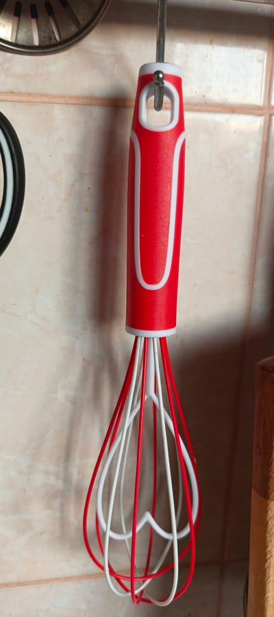 A red and white whisk hangs from a hook against a tiled wall. The whisk has a red plastic handle with white accents and a metal loop for hanging. The whisking wires are a mix of red and white. To the left of the whisk, a portion of a stainless steel strainer is visible. The tiled wall has a light beige color with a subtle pattern. To the right of the whisk, a section of a wooden object, possibly a utensil or part of a cabinet, is visible. The lighting suggests an indoor setting, likely a kitchen, with no indication of the time of day or weather. There is no visible text in the image. The scene is static, with the whisk simply hanging.