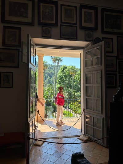 View through a large open window in a room with many framed pictures on the wall, leading to a balcony with a woman in a pink top and tan pants standing on it. Beyond the balcony, there are trees and a building. The image captures part of the Robert Graves House in Bunyola, Spain.