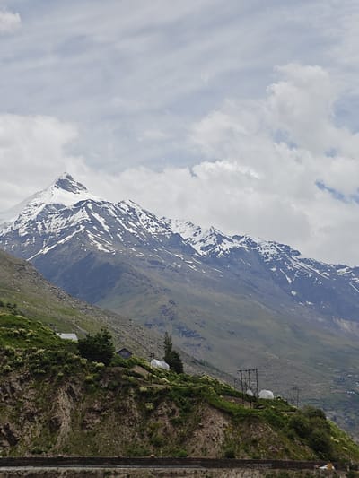 This is a vertical, medium-shot photograph of a snow-capped mountain range under a cloudy sky. The foreground features a steep, rocky, and green hillside with a few small buildings and some vegetation. The middle ground is dominated by the majestic mountain range, with its peaks covered in snow and its slopes showing patches of snow and dark rock. The background consists of a hazy, distant landscape with more mountains and possibly a village. The sky is filled with light gray clouds, hinting at overcast weather. The overall scene depicts a rugged, natural mountain landscape in a remote location, likely in the Himalayas, given the presence of snow-capped peaks and the style of the small structures on the hillside.