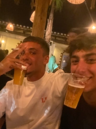 This image depicts two young adult males at night in what appears to be an outdoor or semi-outdoor bar or restaurant setting, consistent with the architecture and vegetation found in the Aljezur municipality of Portugal. The male in the foreground, positioned left of center, is shown smiling while holding a clear plastic cup filled with an amber liquid, likely beer, to his mouth. His right hand is raised, forming a V-sign gesture, with a cigarette held between his index and middle fingers. He is wearing a white long-sleeved shirt with a red 'L' emblem on the chest. To his right, partially obscured, is another male, also smiling and holding a similar clear plastic cup with amber liquid to his mouth. The background is dark, illuminated by multiple overhead woven light fixtures. Visible environmental elements include a building structure with a tiled roofline and the presence of palm trees.