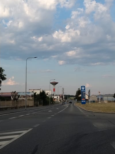This is a vertical, eye-level outdoor shot of a road in Horažďovice, Czechia. The scene appears to be captured during the late afternoon or early evening, with the sun casting long shadows and a soft, diffused light on the landscape. The sky is a mixture of blue and grey clouds, suggesting a partly cloudy day.
The primary focus of the image is a road that curves to the right. White lane markings are visible on the asphalt. To the left of the road, a blue gas station sign with the "ROBIN OIL" logo and prices for different fuels (34.90, 38.50, 35.90, 41.50, 2.19) is prominently displayed. The text "ATRIUM.CZ" and "Area" are also visible on the sign. Further down the road, another sign reads "OTEVÍRÁME C 25.5.2026 PO-SO 9-17", indicating a business opening date and operating hours.
Several buildings are visible in the background, including what appears to be a modern structure with large windows and a water tower. A street lamp stands tall on the left side of the road, illuminating the pathway. A few cars are driving on the road, and there are trees and dry grass along the sides. The overall impression is one of a suburban or commercial area on the outskirts of a town. No people are clearly discernible in the image. The image's orientation is rotated 90 degrees counter-clockwise.