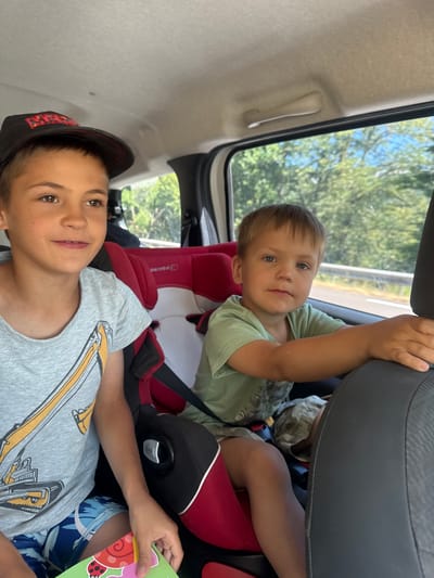 Two young boys are seated in the back of a vehicle, likely on a road trip. The boy on the left, wearing a black baseball cap and a grey t-shirt with a yellow excavator graphic, is looking to his right and smiling. The boy on the right, in a green t-shirt, is looking forward with a neutral expression. He is resting his arm on the back of the car seat in front of him. Both children are secured in car seats. The background visible through the window suggests a natural environment with trees and blue sky. The overall scene depicts a casual moment during travel. The image does not provide specific details about the location in Lavans-lès-Saint-Claude, France, beyond it being a likely route through a rural or suburban area.
