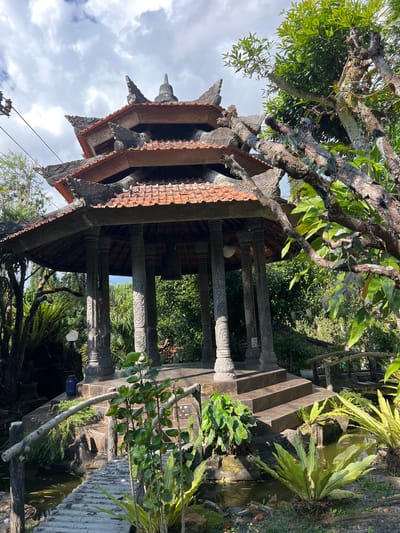 The image depicts an outdoor scene featuring a multi-tiered, octagonal pavilion with a red-tiled roof, characteristic of Balinese architecture. The pavilion is supported by ornate, carved stone pillars. A large bell hangs inside the pavilion. The structure is elevated and accessed by stone steps. A rustic wooden bridge with a simple railing spans a narrow body of water that appears to be a pond or stream, with a small waterfall or rocky edge visible. Lush green vegetation, including ferns and trees, surrounds the pavilion and water feature, suggesting a garden or natural setting. The sky above is partly cloudy with a mix of blue sky and white clouds. There is a blue plastic container and a white lamp post near the pavilion. A power line is visible in the upper left corner. The scene is identified as being in the city of Pupuan, Indonesia. There are no people visible in the image.
