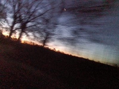 The image depicts a heavily motion-blurred and tilted landscape scene captured during twilight, consistent with either sunrise or sunset. The specified location is Ozark, United States.

The upper portion of the frame is dominated by a sky displaying a gradient from bright orange and yellow along the horizon on the left to dark blue and grey towards the top and right. Streaks of dark and light across the sky suggest cloud formations or significant camera movement during exposure. A small, bright, crescent-shaped object is visible in the upper-mid left quadrant of the sky.

On the left side, several large, dark trees with visible bare branches are silhouetted against the brighter horizon. These trees exhibit severe horizontal motion blur, rendering their forms indistinct.

The lower half of the image consists of a dark, indistinct landmass, likely an uncultivated field or forested area, which forms an undulating horizon line sloping upwards from the left foreground to the right background. No human figures or discernible structures are present within the frame. The overall impression is one of low light and significant dynamic blur.
