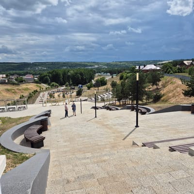 This is a high-angle, full shot image of a public park area in Liski, Russia. The park is situated on a hillside overlooking a valley with a town in the distance. The foreground is dominated by a paved area with stone steps and curved stone benches. Two men are standing on the paved area, one in a pink shirt and dark pants, the other in a plaid shirt and shorts. In the middle ground, there is a playground with various equipment, pathways, and trees. The background shows rolling hills covered in trees and houses. The sky is overcast with dramatic clouds.