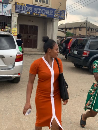 The scene is set on an unpaved urban street in Aba, Abia, Nigeria, identifiable by a Nigerian phone number (080275425637) on a store sign and a vehicle with an Abia state license plate "END 705 CD".
In the foreground, an adult female with dark skin and hair pulled into a bun walks to the right, looking sideways. She wears a short-sleeved, knee-length orange dress with a prominent white vertical stripe down the center, forming a V-neck. She carries a black shoulder bag on her left and holds a clear plastic water bottle in her right hand. To her immediate right, a portion of another adult female is visible, wearing a green floral dress and black slip-on sandals.
In the mid-ground, a silver Toyota Highlander SUV is parked on the left. Directly behind it, a child or young adult in a black top and green patterned bottoms walks away from the camera. On the right side of the street, several vehicles are parked, including a dark SUV (likely a Ford Escape) with the license plate "END 705 CD" and a red SUV behind it, showing a spare tire cover.
The background features a two-story building, on which "MINIKY'S KIDS STORE" is prominently displayed in blue text, advertising "All About Babies" and listing products such as diapers, toys, cosmetics, and clothes. Further in the background, another multi-story building is visible under an overcast sky.