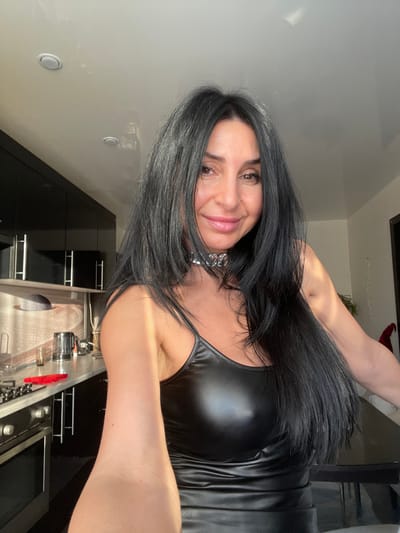 A woman with long, dark hair, wearing a shiny black top and a silver chain necklace, smiles at the camera. She stands in a modern kitchen with black cabinets and stainless steel appliances. A portion of a table or countertop is visible in the background. The image is taken at an angle, giving a tilted perspective.