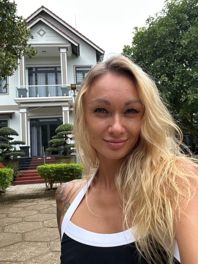 A blonde woman with blue eyes is standing in front of a two-story house in Thôn 2, Vietnam. The house is white with a red tiled roof and has a balcony on the second floor. There are trees and bushes around the house, and the ground is paved with stone. The woman is wearing a black and white tank top and has tattoos on her arm. She is smiling and looking at the camera. The sky is overcast.