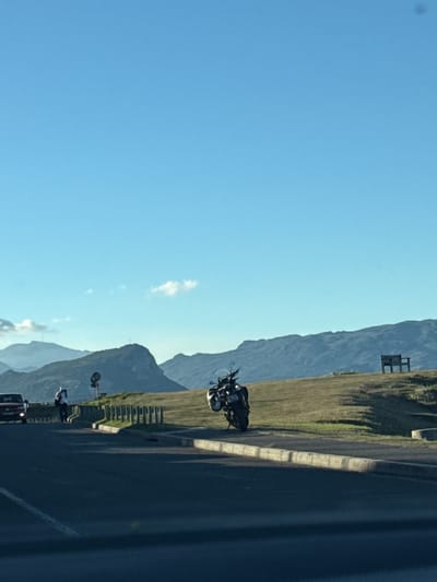 The image shows a landscape in Simon's Town, South Africa. The sky is clear blue with a few scattered clouds. In the midground, there are mountains covered with vegetation. A dark-colored motorcycle is parked on the side of a paved road, with a guardrail separating the road from the grassy mountain slope. Further down the road, a person appears to be standing near a vehicle. The image is oriented vertically, and the elements are positioned to the side. A structure can be seen on top of a higher part of the mountain.