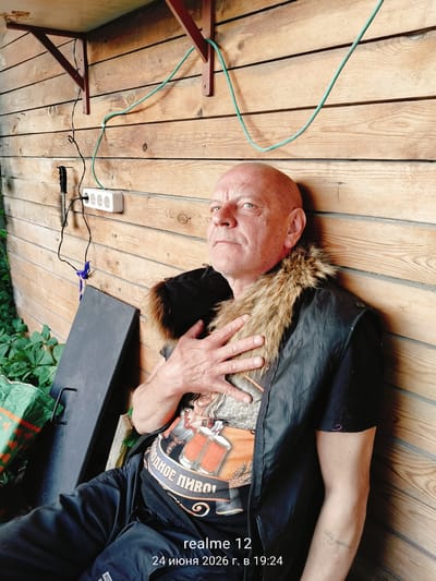 In a sun-drenched, open-air wooden structure, likely a porch or shed, a bald, middle-aged Caucasian man with fair skin and blue eyes is captured in a seated pose. He wears a black vest with a fluffy fur collar over a t-shirt adorned with a graphic of two beer mugs. His left hand rests on his chest, and his gaze is directed upwards and to the left, with a slightly furrowed brow. The wall behind him is constructed of horizontal wooden planks. Electrical conduits and a power strip with three sockets are visible on the wall, along with some hanging wires and a dark, flat object leaning against the wall. Green vines grow lushly to the left of the structure, partially obscuring a dark object that appears to be a bag. The overall impression is one of casual relaxation in an outdoor, utilitarian space. The date stamp indicates the photo was taken on June 24, 2026, at 19:24.