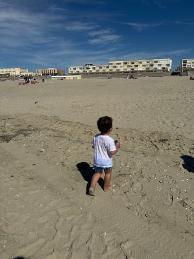 A young child wearing a white t-shirt and blue patterned swim trunks stands on a sandy beach in Berck, France, facing away from the camera. The child's shadow is cast to their right. In the background, several people are visible on the beach, some sitting and others standing. Further back, a row of apartment buildings and colorful beach huts line the coast. The sky is blue with scattered white clouds. The sand is textured with ripples and scattered shells and debris.