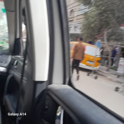 A view from inside a car. Through the car window, a street in Nuseirat, Palestinian Territories, is visible. A person with brown clothing is walking next to a yellow and white cart. Other indistinct buildings and trees are visible in the background. The car's interior is in the foreground, including the dashboard and rearview mirror. The words "Galaxy A14" are written in the bottom right corner.