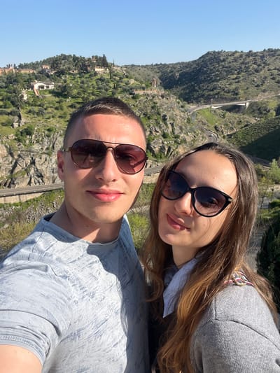 A selfie-style photograph captures two individuals, a male and a female, in the foreground against a panoramic backdrop of the Tagus River valley near Toledo, Spain. The specific location is consistent with views from the Mirador del Valle, overlooking the river gorge and surrounding hills.
The male, positioned to the left, has short dark hair and wears brown aviator-style sunglasses with the "Guess" brand visible on the left temple. He is dressed in a light grey short-sleeved t-shirt featuring a subtle textured or patterned print. He maintains a slight smile and looks directly towards the camera.
The female, positioned to the right, has long brown hair and wears dark cat-eye style sunglasses. She is wearing a grey knit sweater over a light blue collared shirt. A multi-colored, possibly striped, decorative pin or brooch is attached to her right shoulder. She also displays a slight smile and looks towards the camera.
The background consists of a clear blue sky above a rugged, green landscape. Multiple hills are visible, covered with trees, shrubs, and exposed rock formations. A winding road descends through the valley, and a modern white concrete bridge, consistent with the Puente de la Cava, spans across a lower section of the valley. Scattered buildings are discernible on the distant hillsides. The scene is brightly illuminated by sunlight, casting shadows on the subjects.