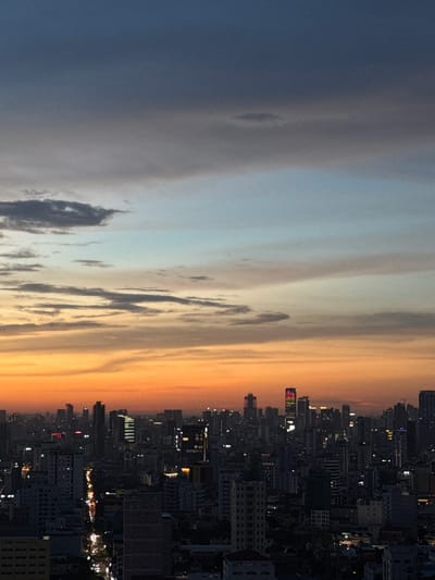 The image depicts a sprawling cityscape at dusk, with a dramatic sky transitioning from a deep blue on the left to a vibrant orange on the right, indicating the end of the day. Wispy clouds stretch across the sky, adding texture and depth to the scene.

The cityscape itself is densely populated with numerous buildings of varying heights, many of which are illuminated, suggesting the early evening hours. The lights from the buildings create a twinkling effect against the darkening sky, highlighting the urban sprawl. Some buildings are lit with colorful lights, adding a festive or commercial element to the view.

The perspective is from a high vantage point, likely an elevated building or viewpoint, offering a panoramic view of the city. The composition emphasizes the contrast between the natural beauty of the sky and the man-made structures of the city. The overall mood is one of urban dynamism transitioning into the calm of the evening. No visible text is present in the image. The location context is Khan Mean Chey, Cambodia, which would be consistent with a dense urban environment in Southeast Asia experiencing a sunset.