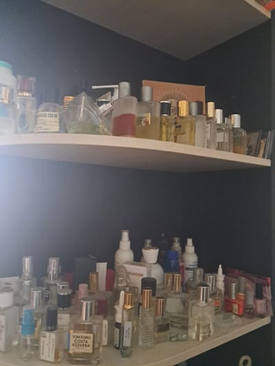 A top-down, slightly angled, medium shot captures two shelves filled with various perfume bottles and other cosmetic products. The shelves are made of a light-colored, smooth material and are positioned against a dark background.
The top shelf displays a collection of perfume bottles with a variety of shapes and sizes. Some bottles are clear glass, revealing different colored liquids inside, while others are opaque. Notable bottles include those with silver and gold caps, and some with intricate designs. A few smaller, cylindrical bottles are also visible. In the background, a partially obscured orange artwork with a radiating design is present.
The bottom shelf is also densely packed with perfume bottles. Here, the bottles are more varied in color and design. Some have metallic caps, while others have plastic caps in shades of blue, pink, and white. A prominent bottle in the center of this shelf has "TOM FORD COSTA AZZURRA" printed on its label. To the right of this, a bottle with a gold cap and a light-colored liquid is visible. Further to the right, nail polish bottles in shades of red, pink, and possibly orange are arranged. A white spray bottle with a blue label is also visible on this shelf. The overall impression is one of an organized, albeit full, collection of personal care products.
The image does not contain any information that could place it in Solnechnogorsk, Russia.