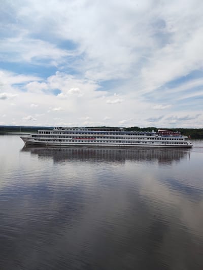 A long, white river cruise ship is sailing on a wide, calm river. The ship has multiple decks and many windows. The reflection of the ship and the sky is visible on the water's surface. The background shows a dark green, dense forest along the riverbank. The sky is partly cloudy with patches of blue. The scene is set in Shchetinino, Russia. There are no people visible in the image.