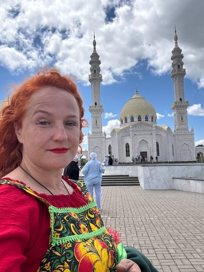 This is a vertical, outdoor, selfie-style photo taken on a bright, cloudy day. The main subject is a woman with vibrant red hair and bright red lipstick, looking directly at the camera. She is wearing a red top with a black string strap and a colorful, patterned overlay featuring yellow, green, and red floral designs, reminiscent of traditional Russian folk art.

In the background, a large, white mosque with a golden dome and tall minarets is visible. The architecture appears grand and ornate. People, some wearing white head coverings, are seen on steps leading up to the mosque entrance, suggesting a place of worship or a tourist attraction. The ground is paved with light-colored rectangular bricks. The sky is a vibrant blue with scattered white clouds.

The overall mood conveyed by the woman's direct gaze and makeup is confident and engaging. The location context of Privolzhsky, Russia, combined with the traditional folk art pattern on her clothing and the prominent mosque, suggests a cultural or sightseeing visit.