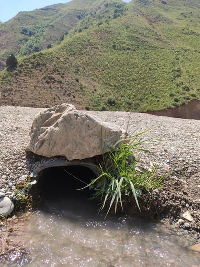 A large, light-colored boulder sits partially covering the entrance of a dark, circular culvert. Green grass grows around the opening of the culvert and a small stream of murky water flows out from it, creating ripples and reflections on the surface. The culvert and stream are situated on a gravelly surface. In the background, a steep, green, mountainous landscape rises under a clear blue sky. There are no people visible in the image. The scene appears to be in a natural, outdoor setting, likely in a rural or mountainous area. The location is identified as Sadika, Tajikistan.