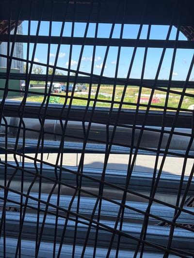 The image shows a view through a dark metal grate, looking out onto an outdoor industrial or agricultural area under a bright blue sky with scattered white clouds. In the background, several trucks and what appear to be silos or storage tanks are visible. The landscape includes some green vegetation and patches of lighter colored ground. The grate itself is made of thick, dark wires, some of which are bent. In the foreground and middle ground, there are horizontal metal structures, possibly part of the building the photo is taken from or stacked materials. One prominent object in the middle ground is a large, metallic cylindrical object. The scene appears to be located in Rąbczyn, Poland, suggesting an industrial or agricultural setting within that municipality.